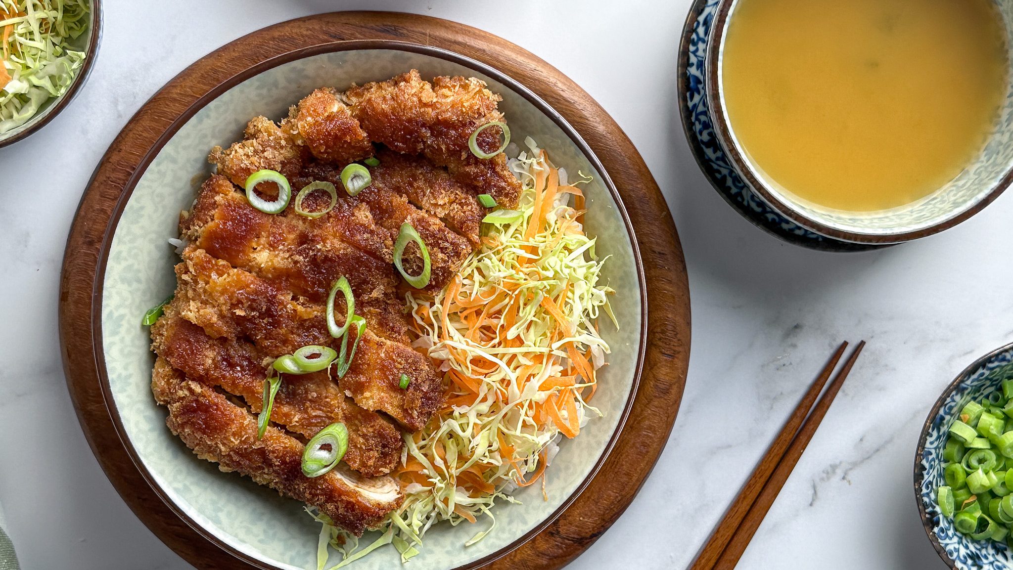 A bowl of shredded cabbage with bread crumbed fried meat, with sauce. A pair of chopsticks and a small bowl of soup.