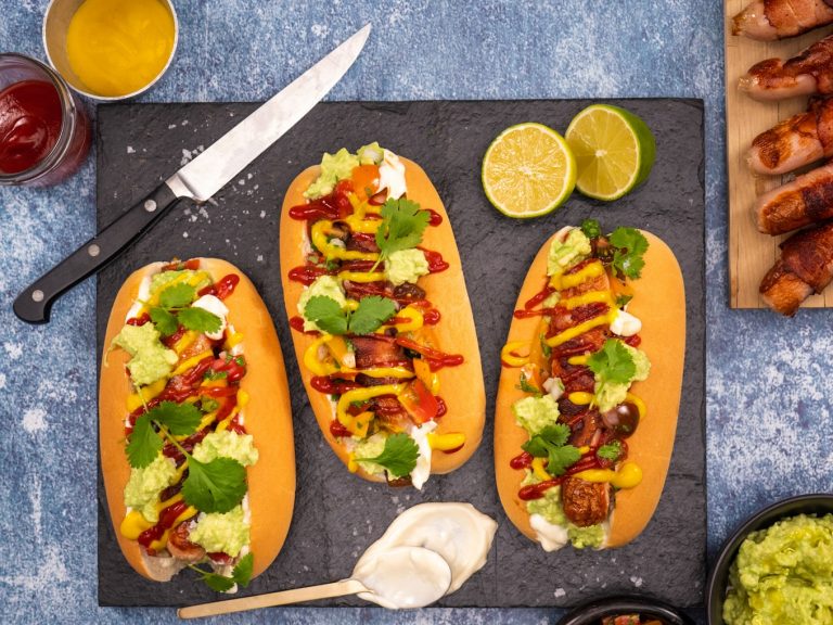 Three loaded hotdogs dressed with guacamole, mustard, tomato sauce and herbs on slate