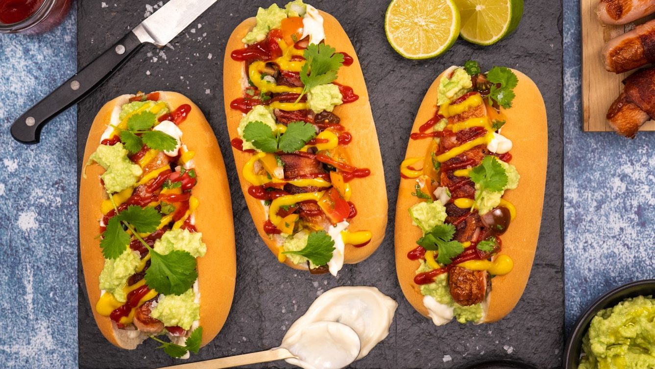 Three loaded hotdogs dressed with guacamole, mustard, tomato sauce and herbs on slate