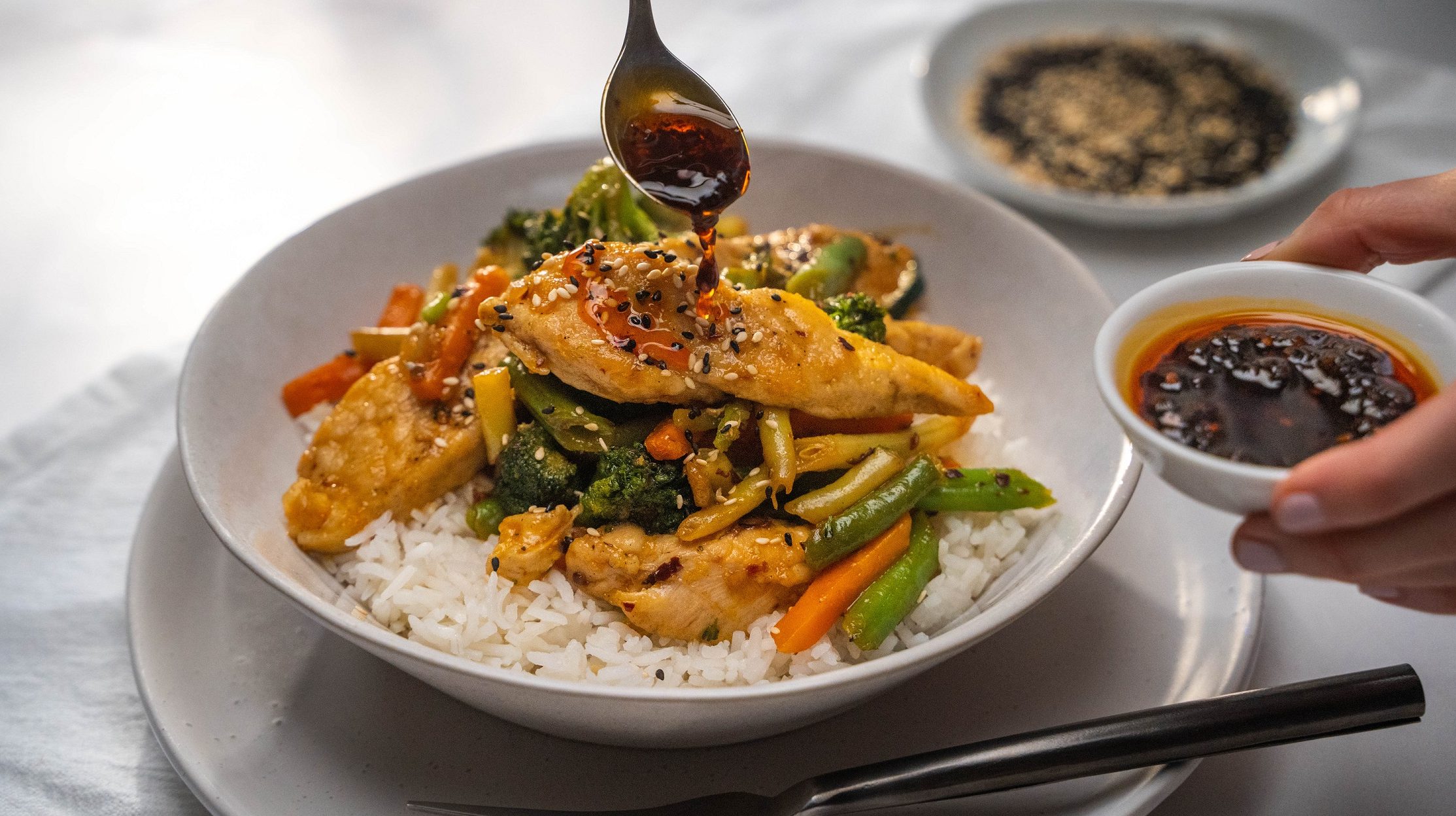 A bowl of chicken and vege stir-fry on white rice, pouring chill sauce with a spoon.