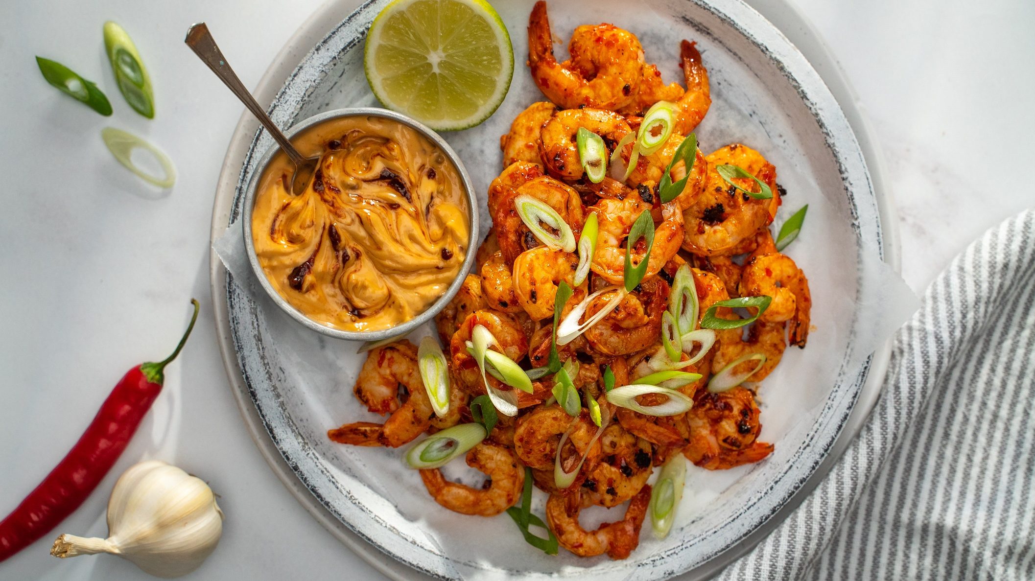 A plate of chilli prawns with a pot of creamy sauce and lime half.