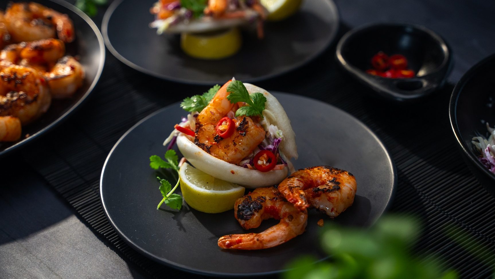 A red prawn and herb filled bao bun on a black plate with more prawns.