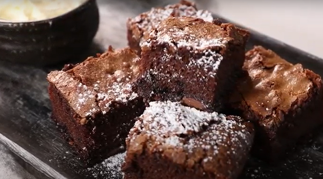 A few chunks of chocolate brownies stack on black plate dusted with icing sugar and a pot of cream in the back