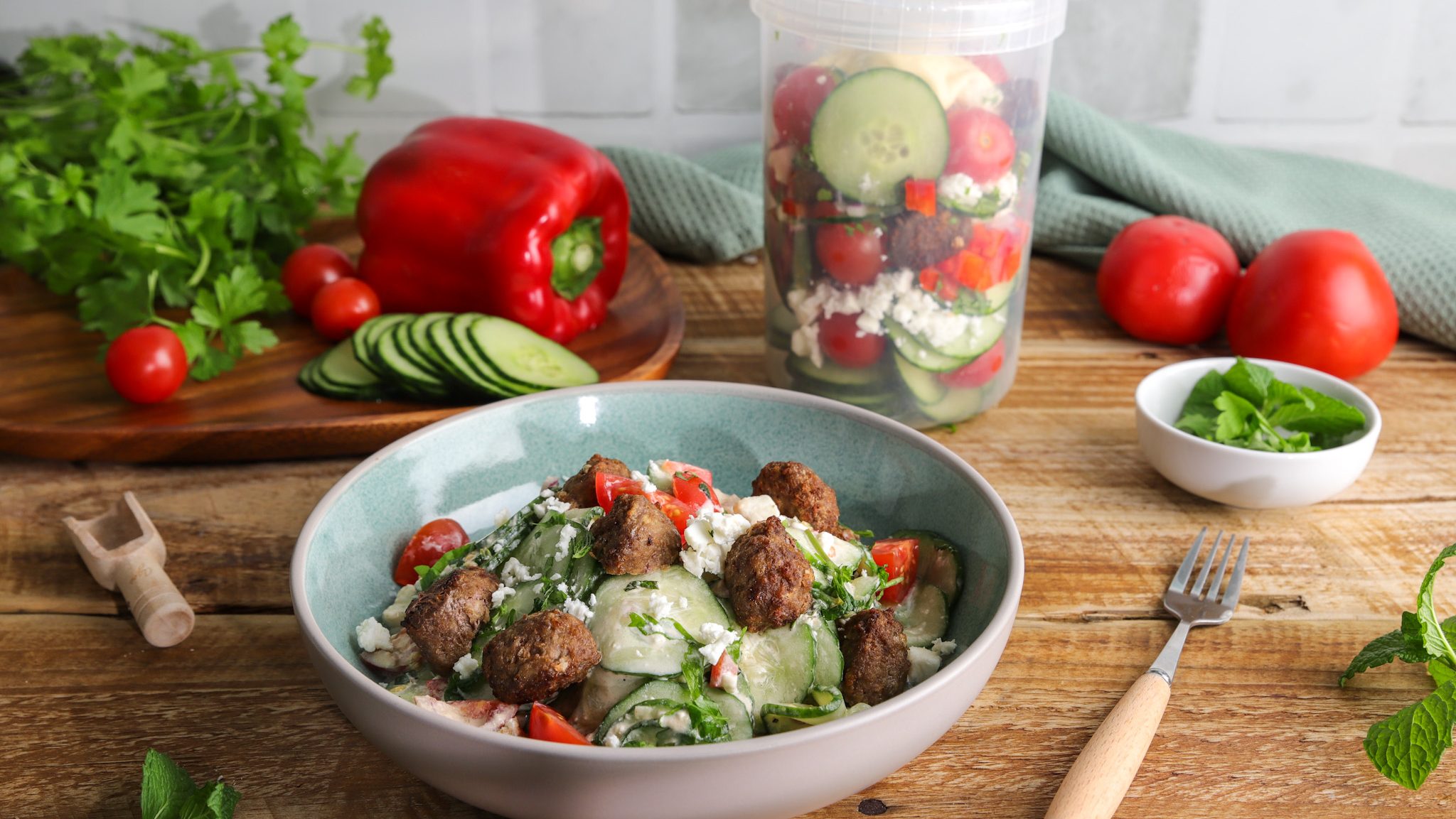 A bowl and a plastic container of meatball salad on a table with raw vegetables.