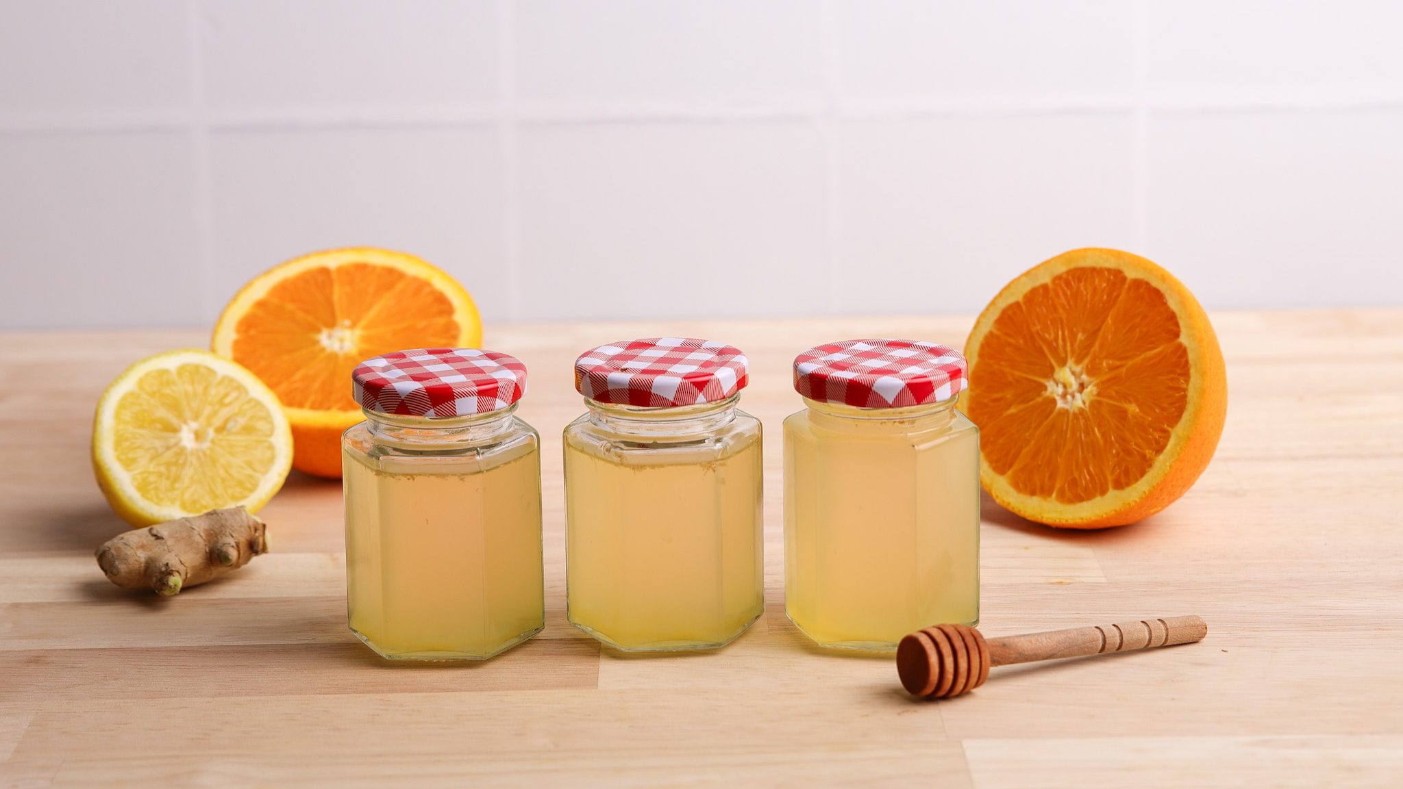 Three jars of pale-yellow liquid, ginger, lemon & orange halves and wooden honey dipping stick on kitchen bench.