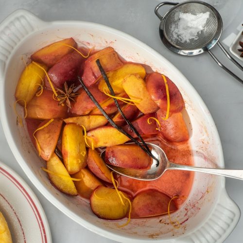 A white oven dish full of orange red coloured baked fruit wedges with a sliver fork, spice on small plate and a tea strainer at side.