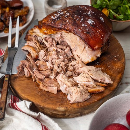 Cooked pork joint shredded on wooden board with knife and fork, apples in front