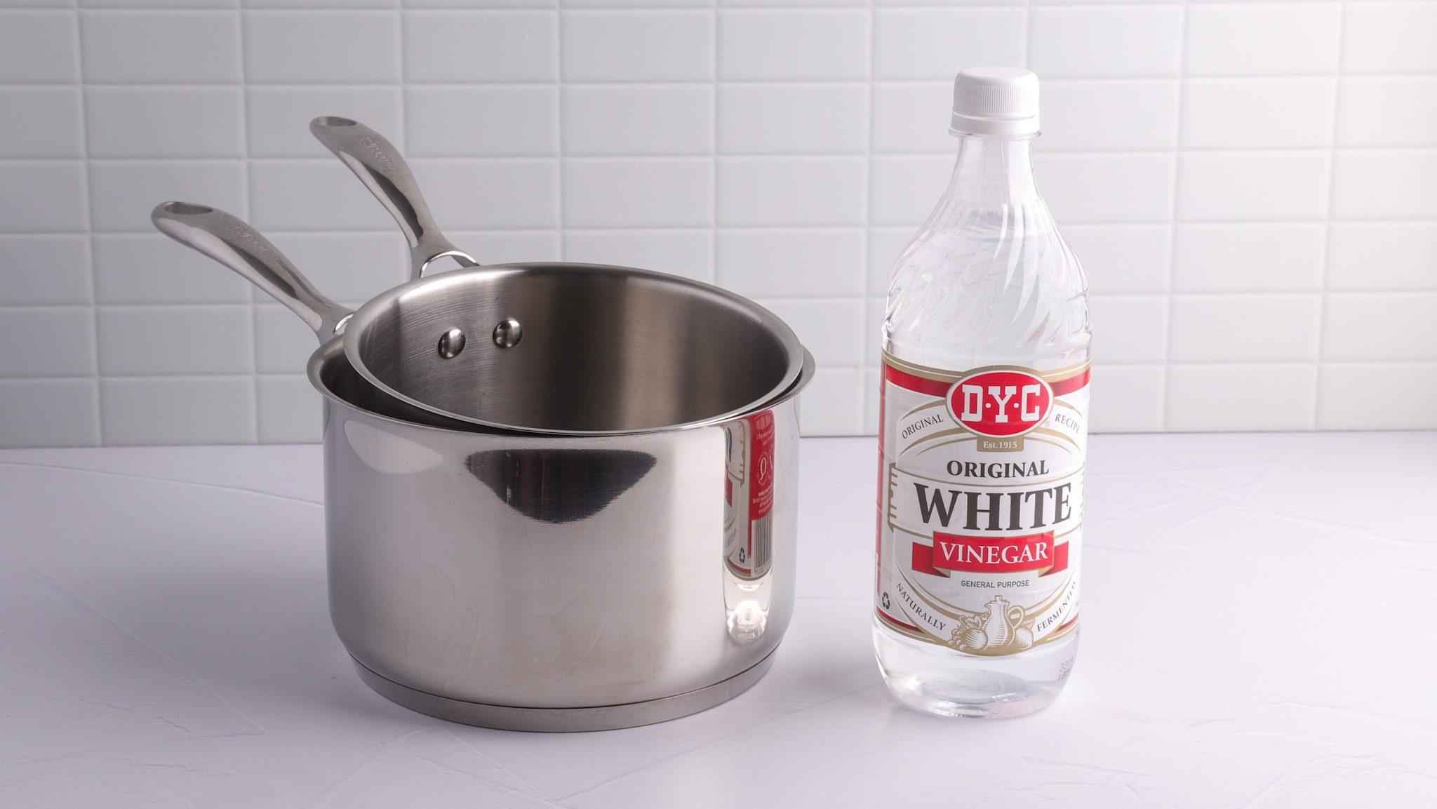 Two stainless steel pots and a bottle of vinegar on white surface.