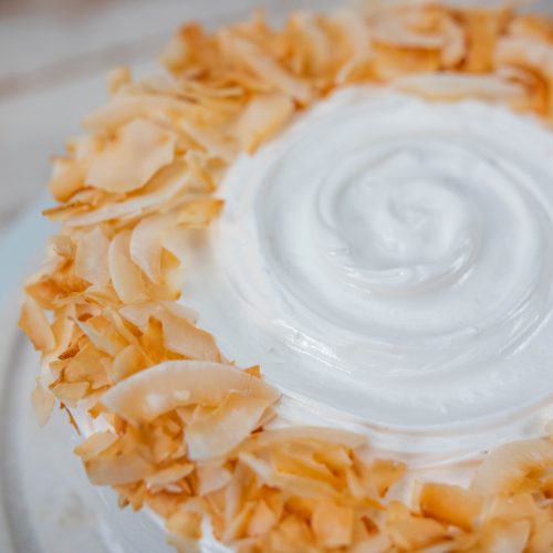Coconut cake in a close up top view, focusing on the icing with a swirl pattern in the center and toasted coconut flakes liberally sprinkled on the edge.