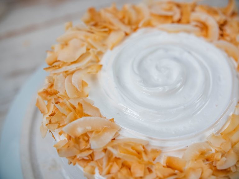 Coconut cake in a close up top view, focusing on the icing with a swirl pattern in the center and toasted coconut flakes liberally sprinkled on the edge.