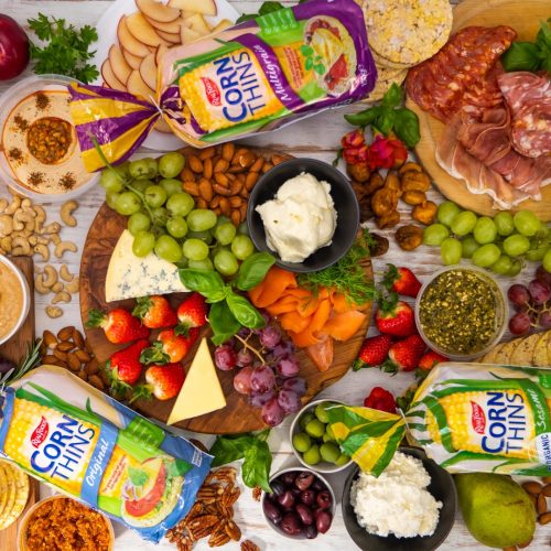 Top view of a grazing platter with a lot of fruits cheese,, 3 bags of corn thins among lots of food.