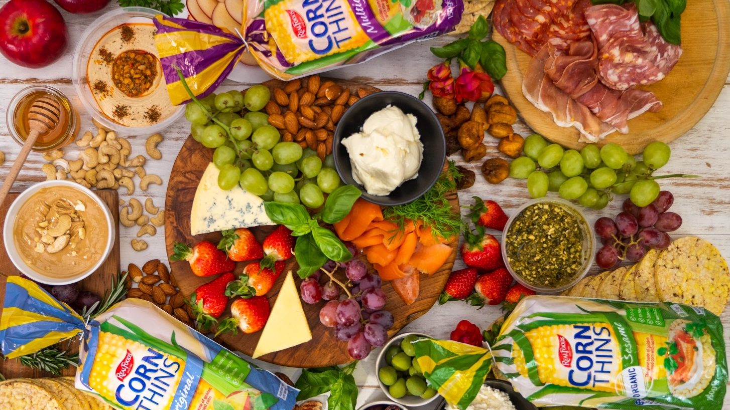Top view of a grazing platter with a lot of fruits cheese,, 3 bags of corn thins among lots of food.