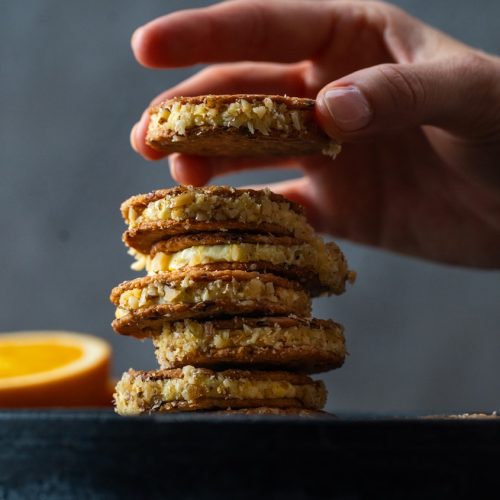 Filled round crackers stack on black slate next to cut orange, a hand is putting another cracker sandwich on top.