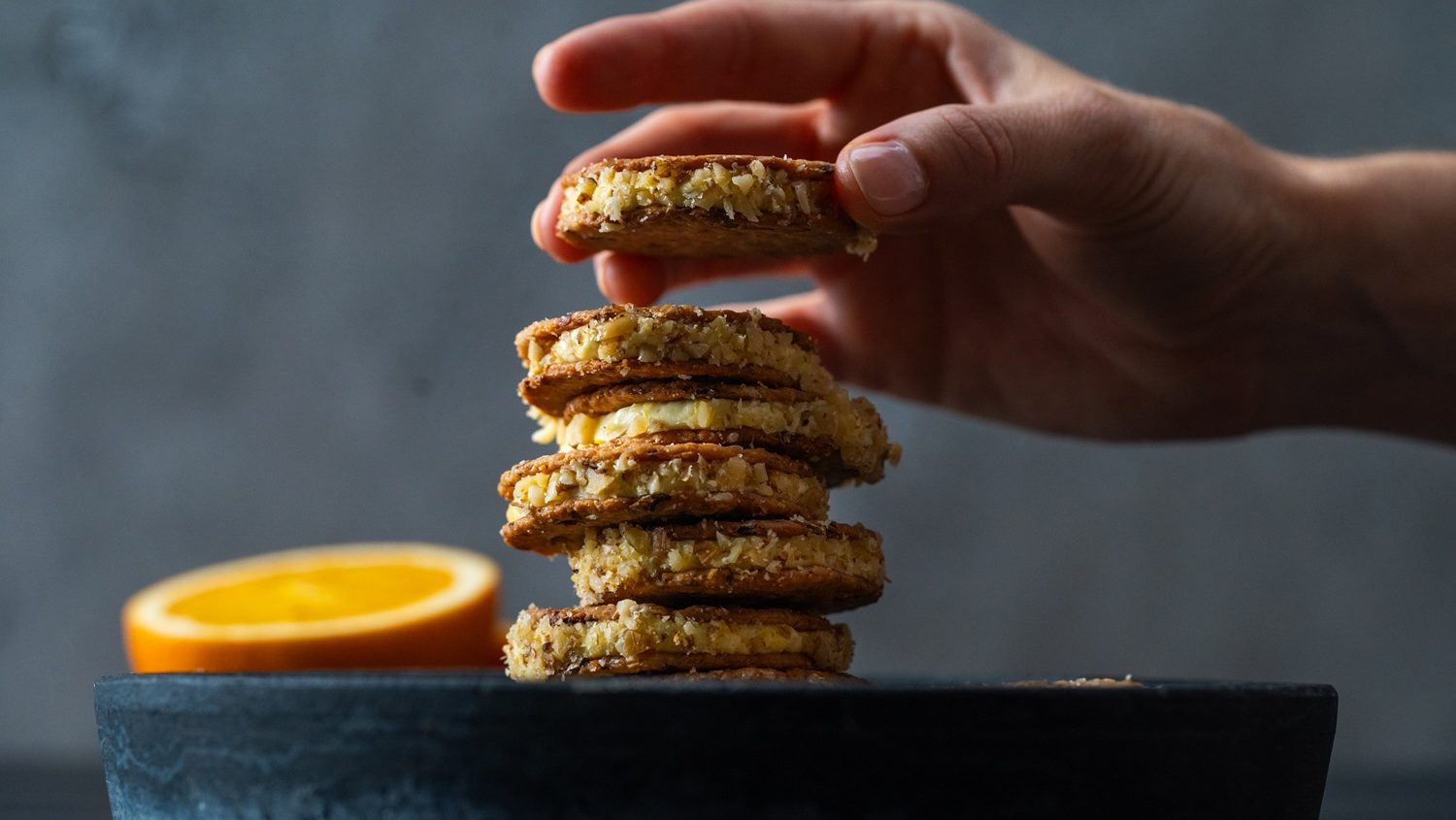 Filled round crackers stack on black slate next to cut orange, a hand is putting another cracker sandwich on top.