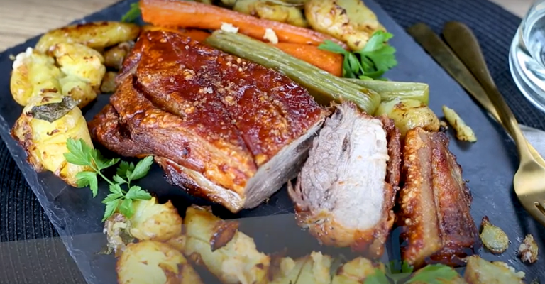 Cooked pork sliced surrounded by potatoes and vegetables on black slate.
