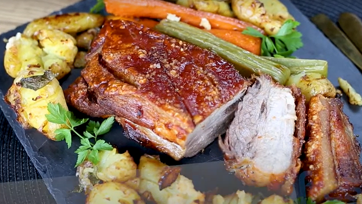 Cooked pork sliced surrounded by potatoes and vegetables on black slate.