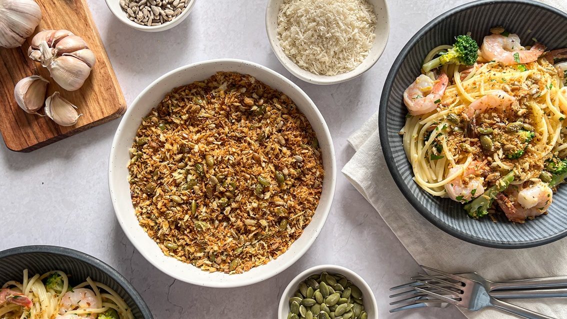 Browned crumbs in a bowl in centre with small bowls of garlic, seeds and spaghetti around it.