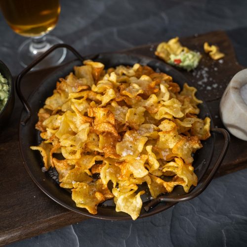 A platter of brown curled pasta chips in centre, on left side a bowl of green aioli , on right side a small pot of salt. A glass of beer in back.