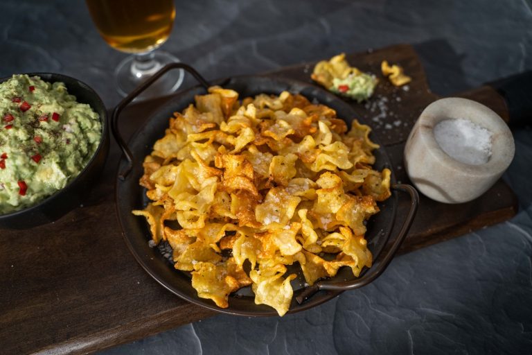 A platter of brown curled pasta chips in centre, on left side a bowl of green aioli , on right side a small pot of salt. A glass of beer in back.