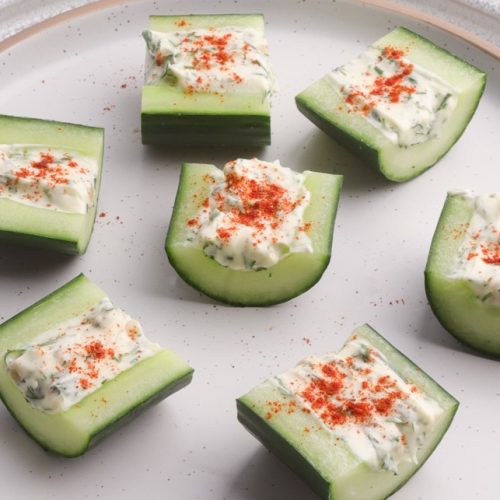 Seven pieces of cream filled cucumber chunks topped with red powder on a plate.
