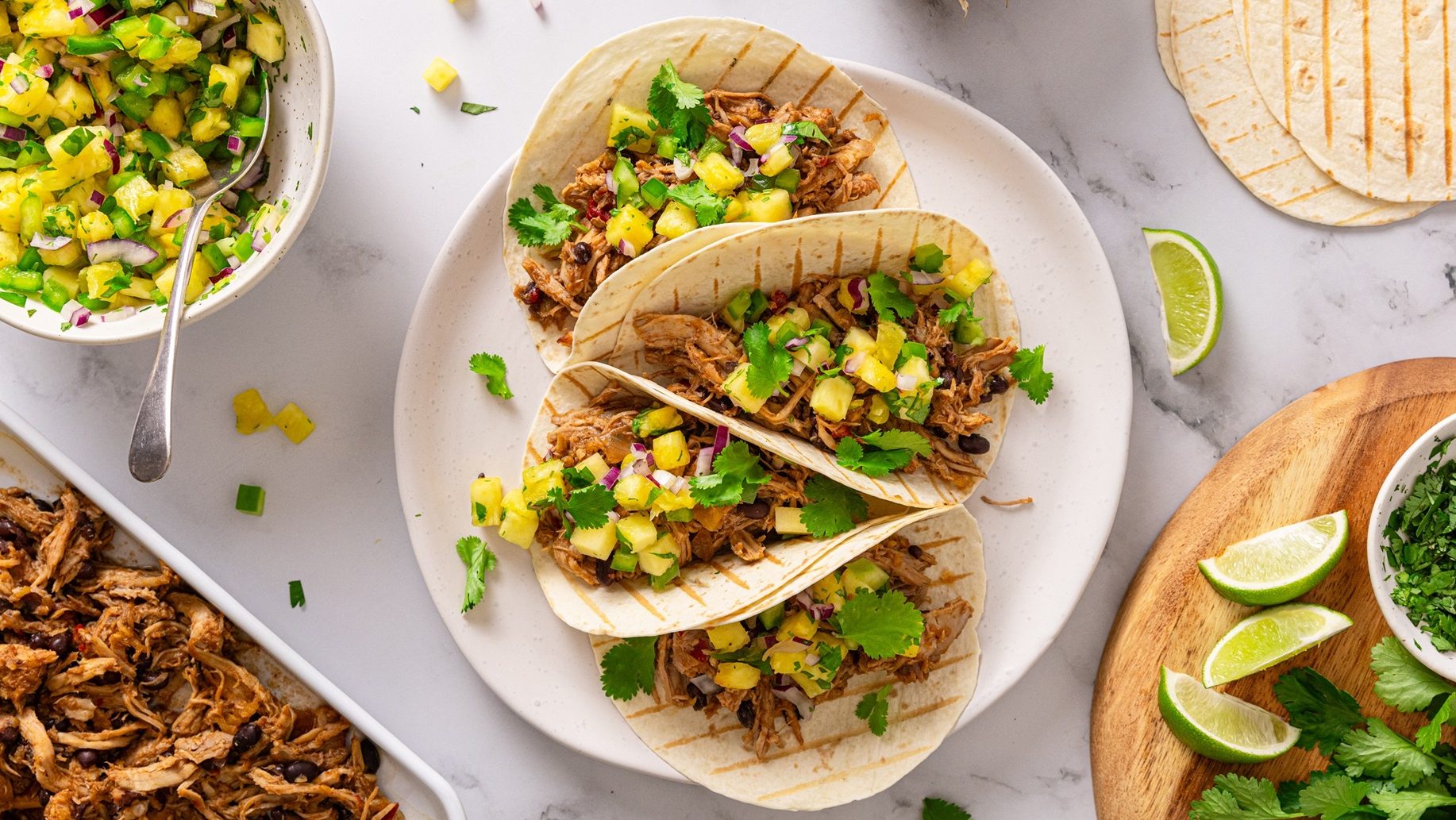 Four tacos filled with brown, yellow and green food on white plate. Surrounded by ingredients.