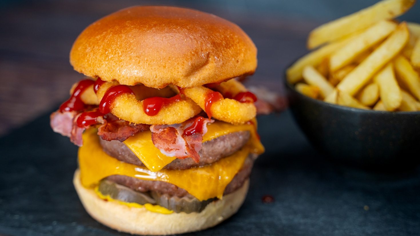 Loaded double cheeseburger on wooden board with a bowl of chips.