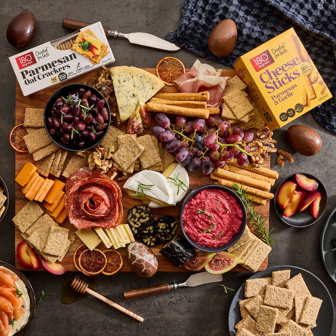A grazing platter of cheese, dips, and crackers, with a packet of 180 Degrees Cheese Sticks and a packet of 180 Degrees Parmesan Oat Crackers laid on the board.