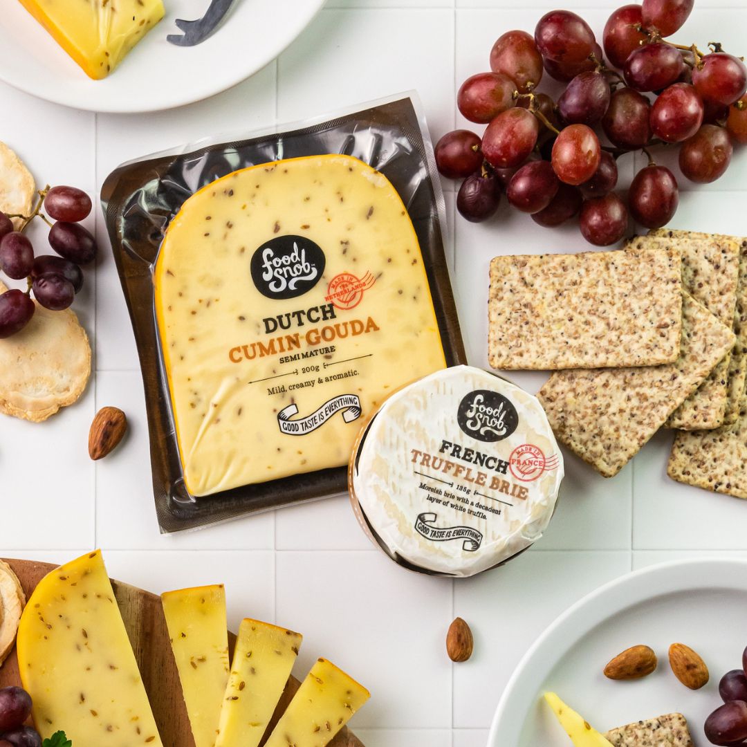 Packets of Food Snob French Truffle Brie and Food Snob Dutch Cumin Gouda surrounded by crackers, grapes, and slices of cheese
