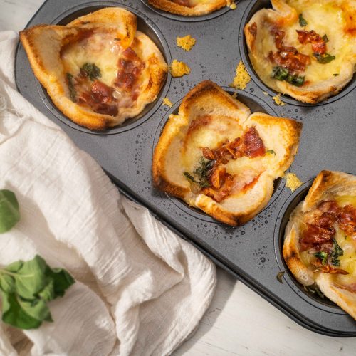 A six-hole muffin tin full of bread cup with pizza fillings and herb.