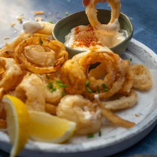 Plate of fried onion and squid rings with lemon wedges and a pot of dipping sauce.