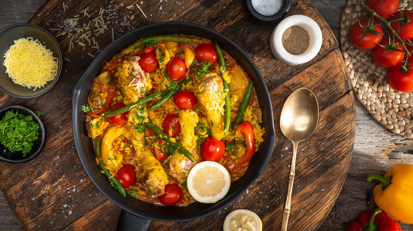 A large skillet with colourful dish, a spoon, small dishes of food and vine tomatoes.