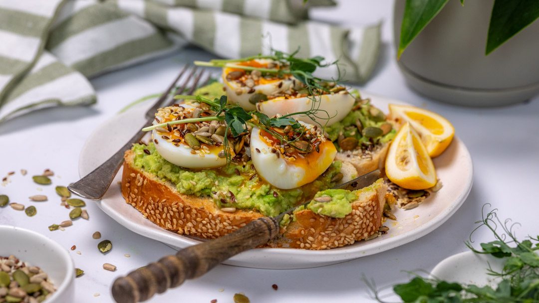 Two pieces of toast topped with smashed avocado and halved boiled eggs with seeds scattered all over.