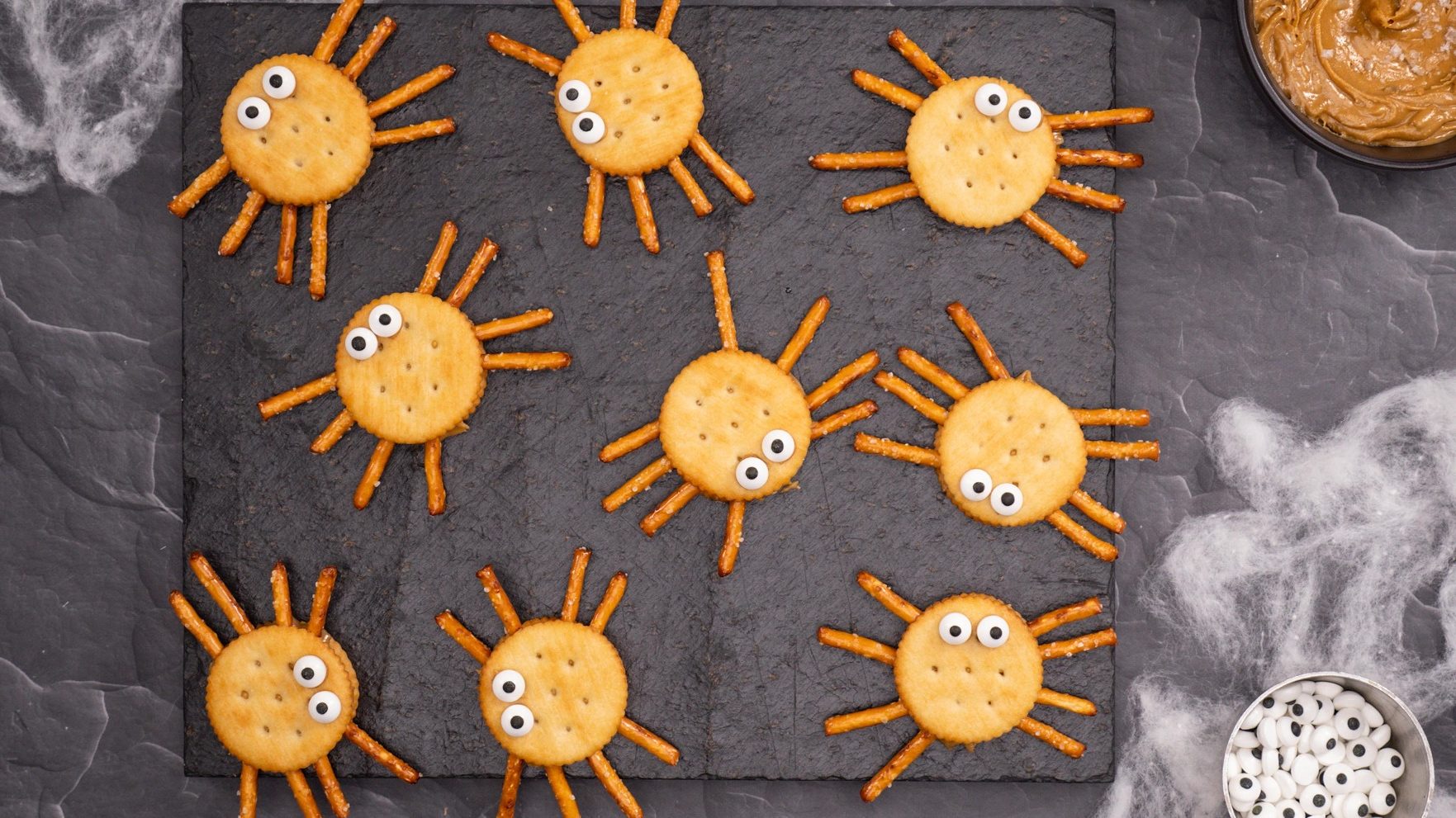Nine cracker sandwich Halloween spiders with pretzel legs and candy eyes on slate