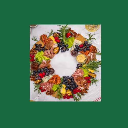 Looking down on an appetising, edible Christmas wreath assembled with berries, charcuterie, avocado, cheese, grapes, stuffed dates, pecans, hummus dip and decorated with fresh sprigs of rosemary.