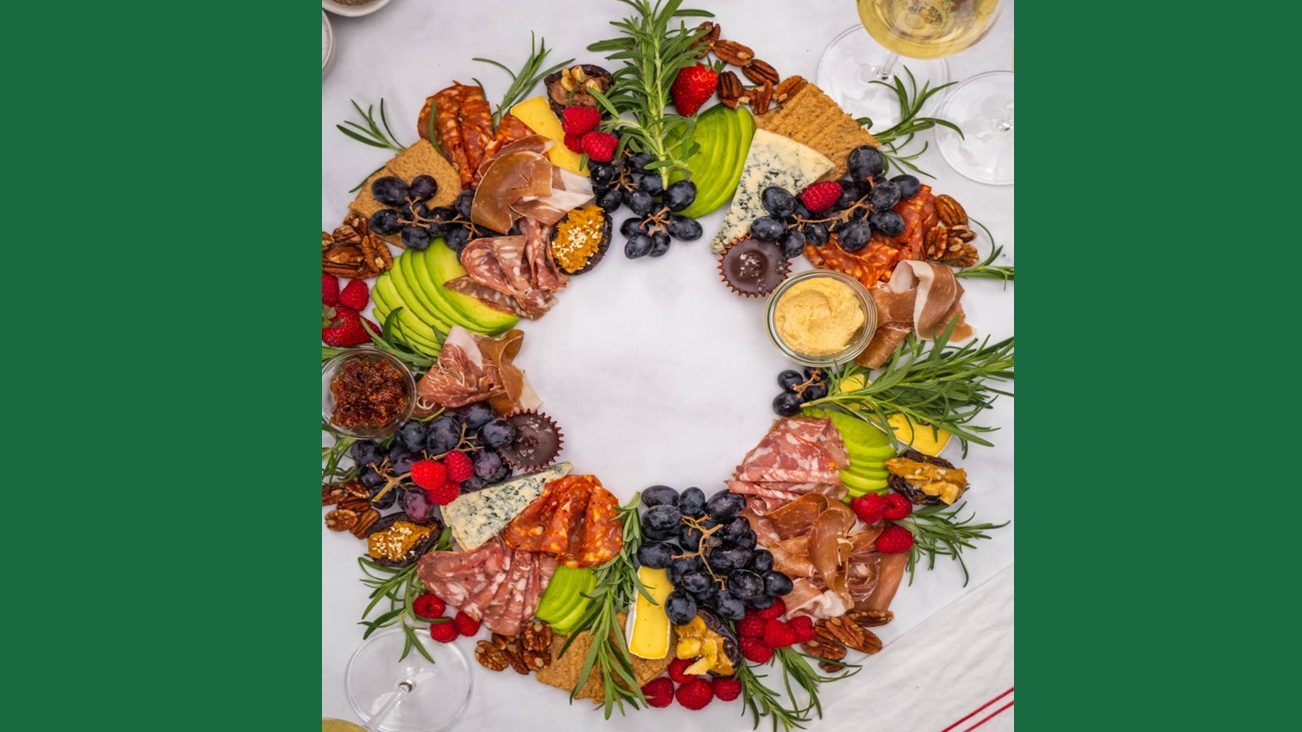 Looking down on an appetising, edible Christmas wreath assembled with berries, charcuterie, avocado, cheese, grapes, stuffed dates, pecans, hummus dip and decorated with fresh sprigs of rosemary.