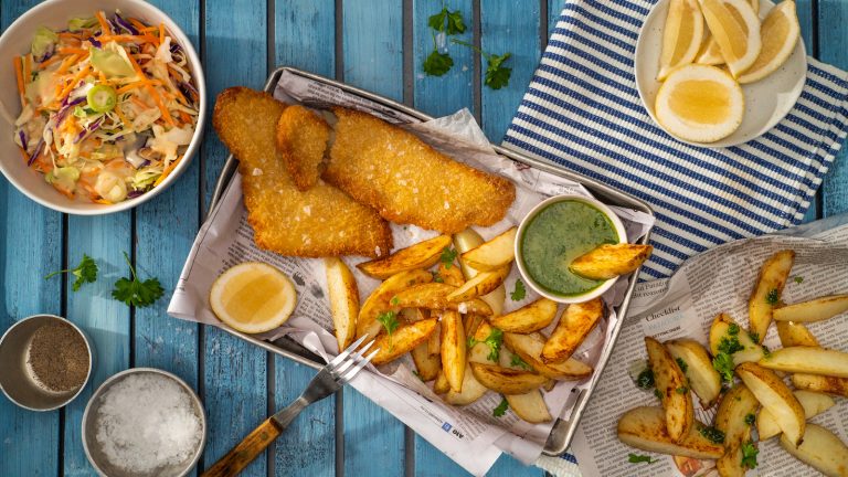 Crumbed fish fillets with potato wedges on a tray, lemon slices, green herb sauce, and a bowl of coleslaw