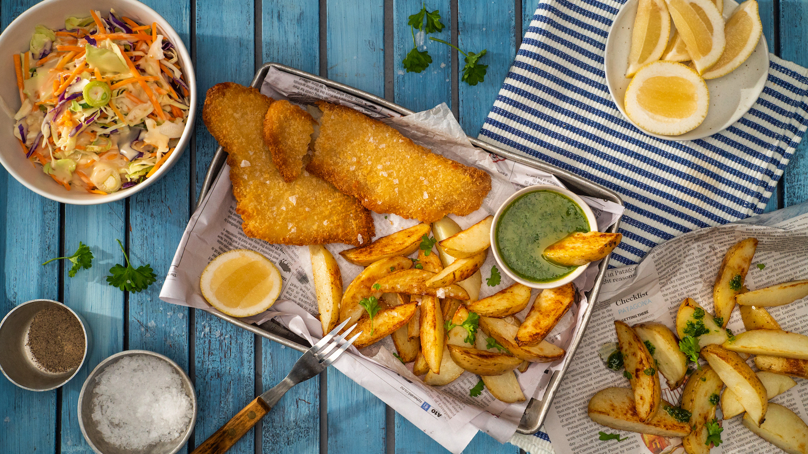 Crumbed fish fillets with potato wedges on a tray, lemon slices, green herb sauce, and a bowl of coleslaw