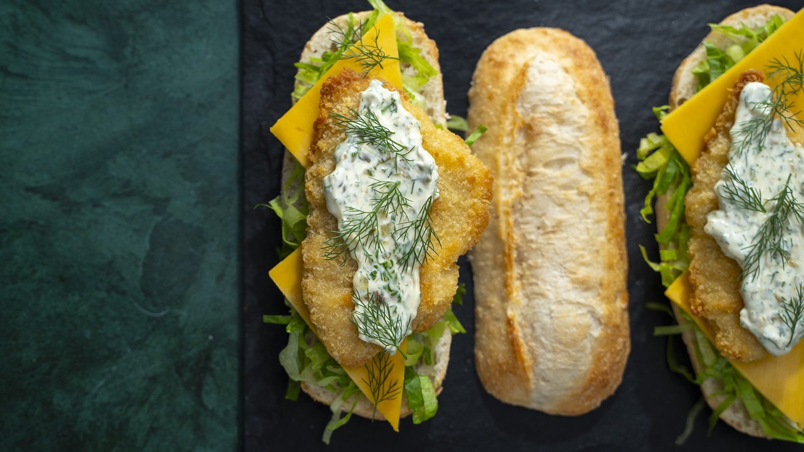 Cheese slices, lettuce, crumbed fish and tartar sauce on an oblong bread with the top half of bread on its side.