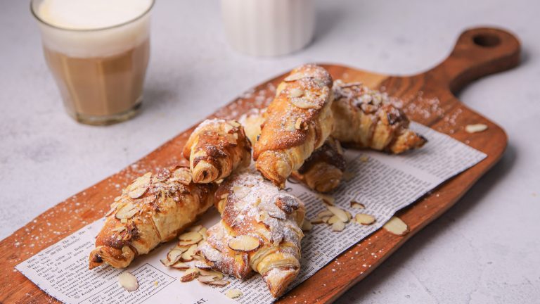 Five almond croissants on a wooden board with a glass of milky coffee behind.