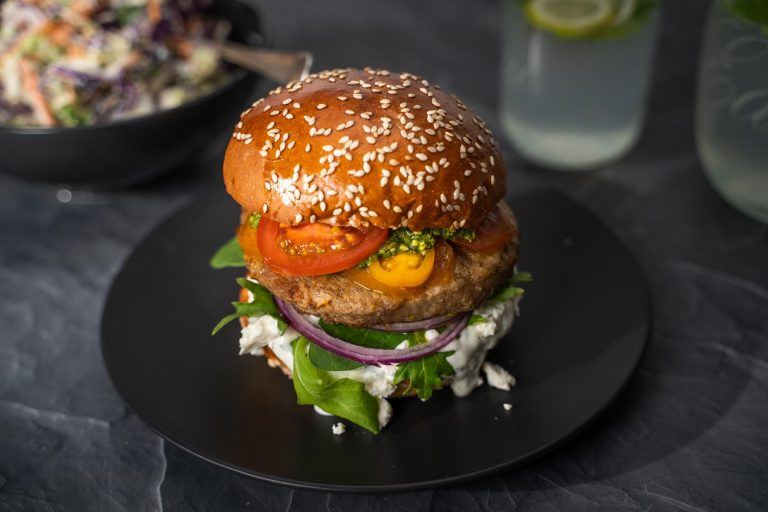 A big burger with green lettuce, white sauce, red onion, pattie, sliced tomatoes in filling on a black round plate. A bowl of coleslaw and glasses of drinks in the back.