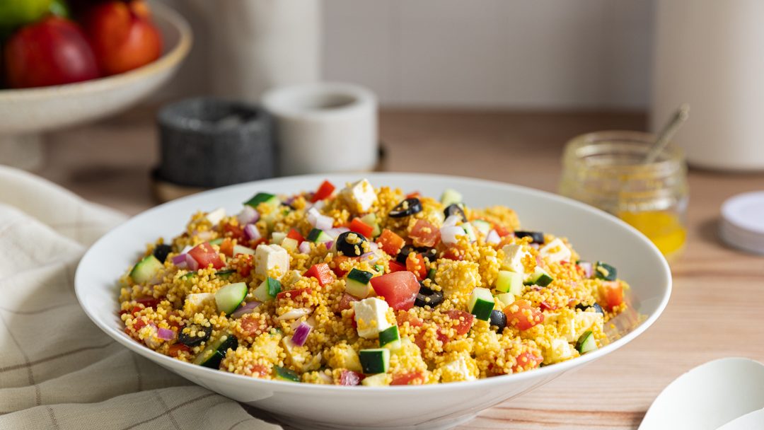 A bowl of yellow couscous with cubes of green, red, white and black foods.