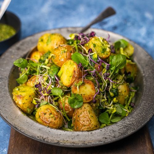 A heap of potato, micro shoots and greens salad on a metal plate with a black bowl of green sauce on blue table.