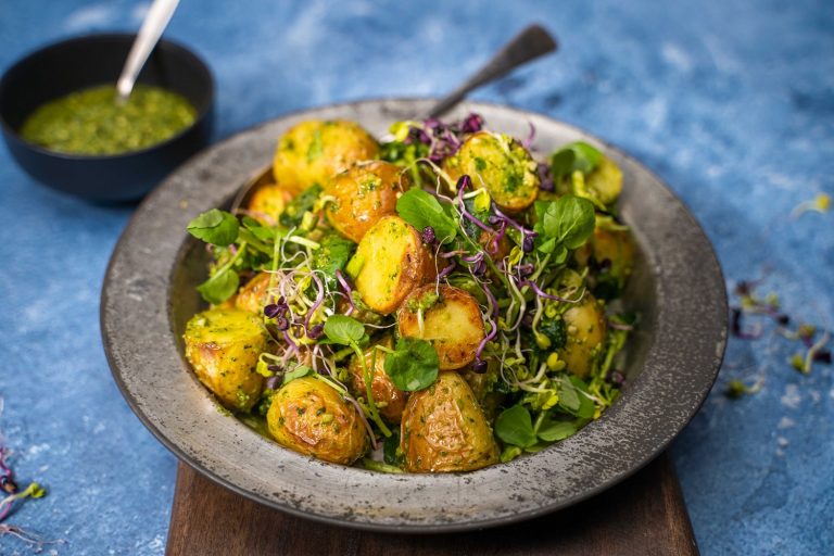 A heap of potato, micro shoots and greens salad on a metal plate with a black bowl of green sauce on blue table.