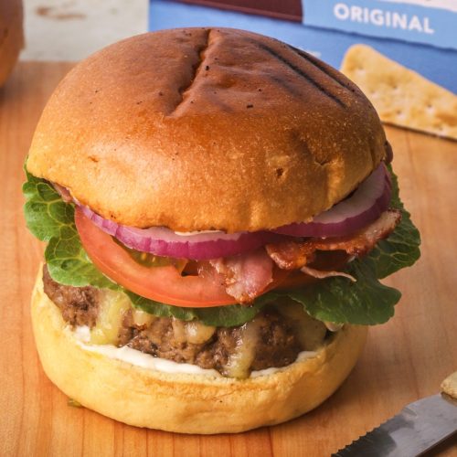A burger with thick beef patty, lettuce, tomato, red onion. A box of crackers at rear.