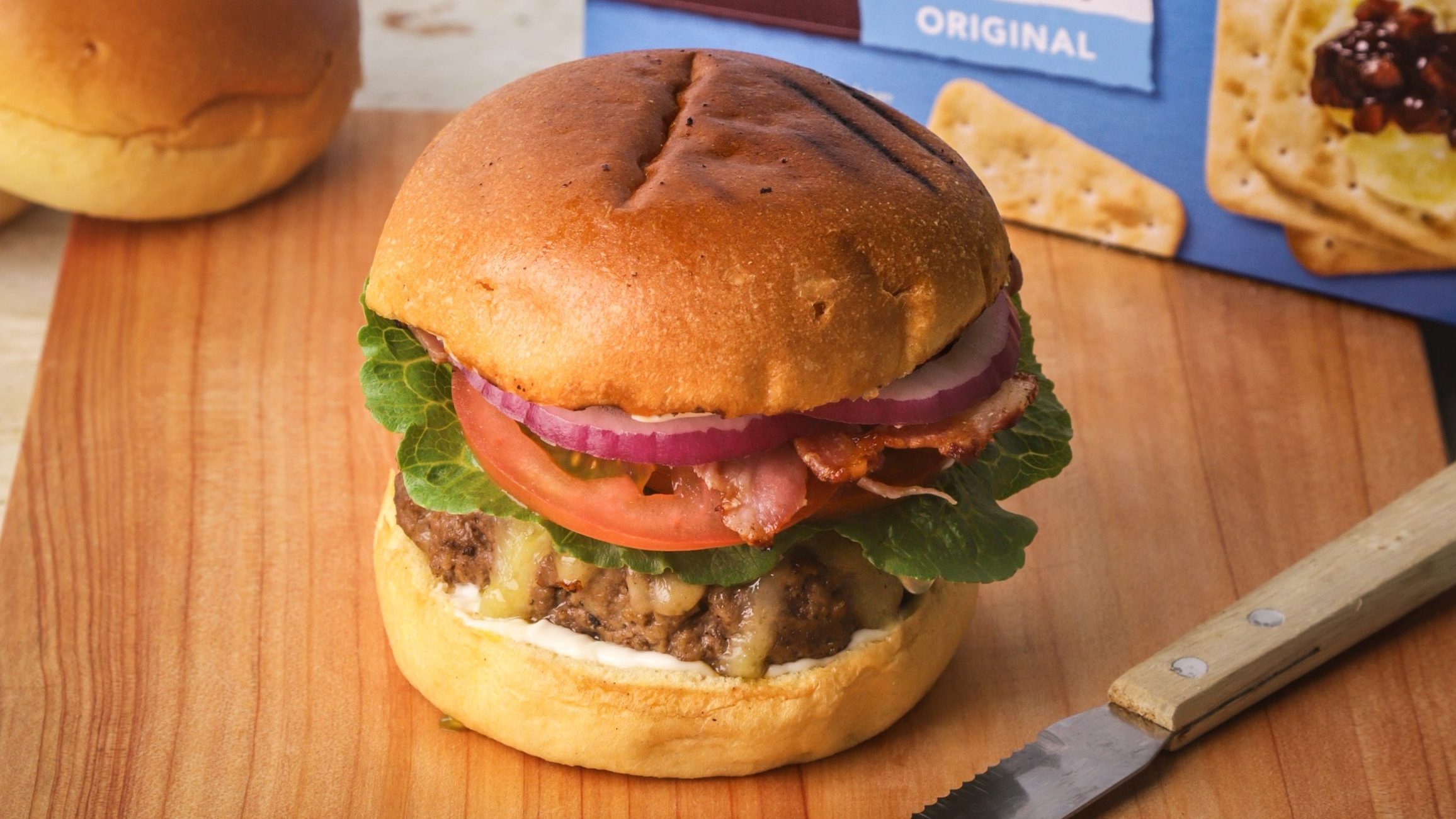 A burger with thick beef patty, lettuce, tomato, red onion. A box of crackers at rear.