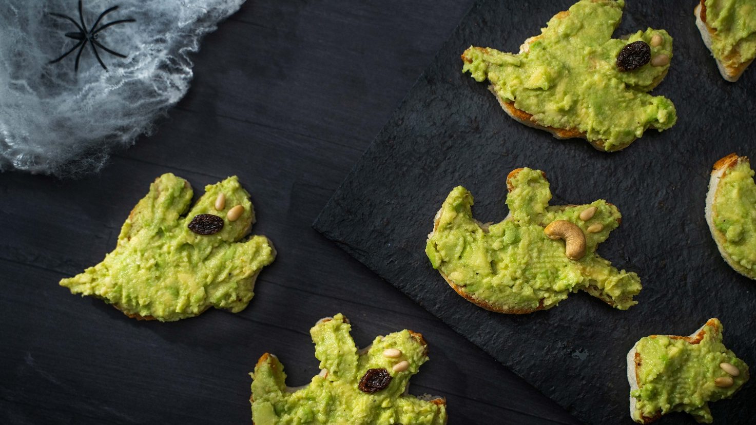 Green mash pasted ghost shaped snacks on black slate with white cotton spider web.