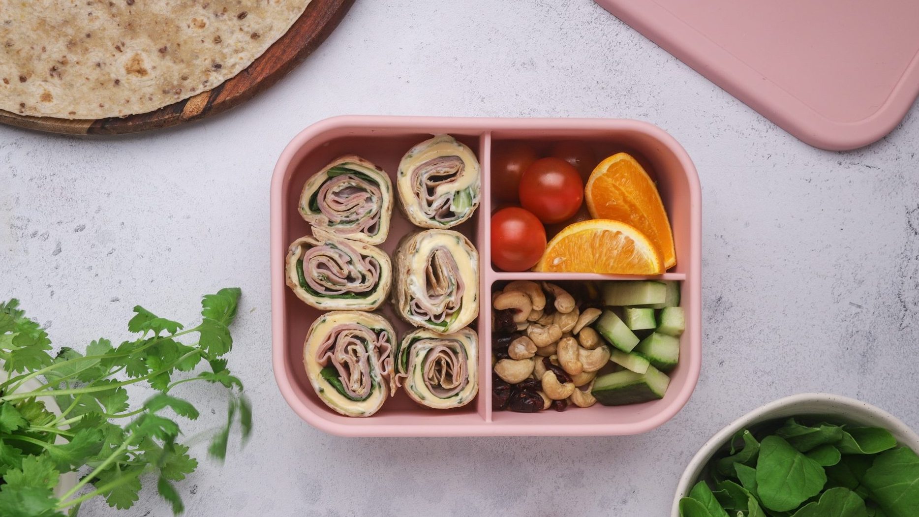 Pink lunch box with six ham pinwheels, mini tomatoes and orange wedges, and vegetable sticks filled.