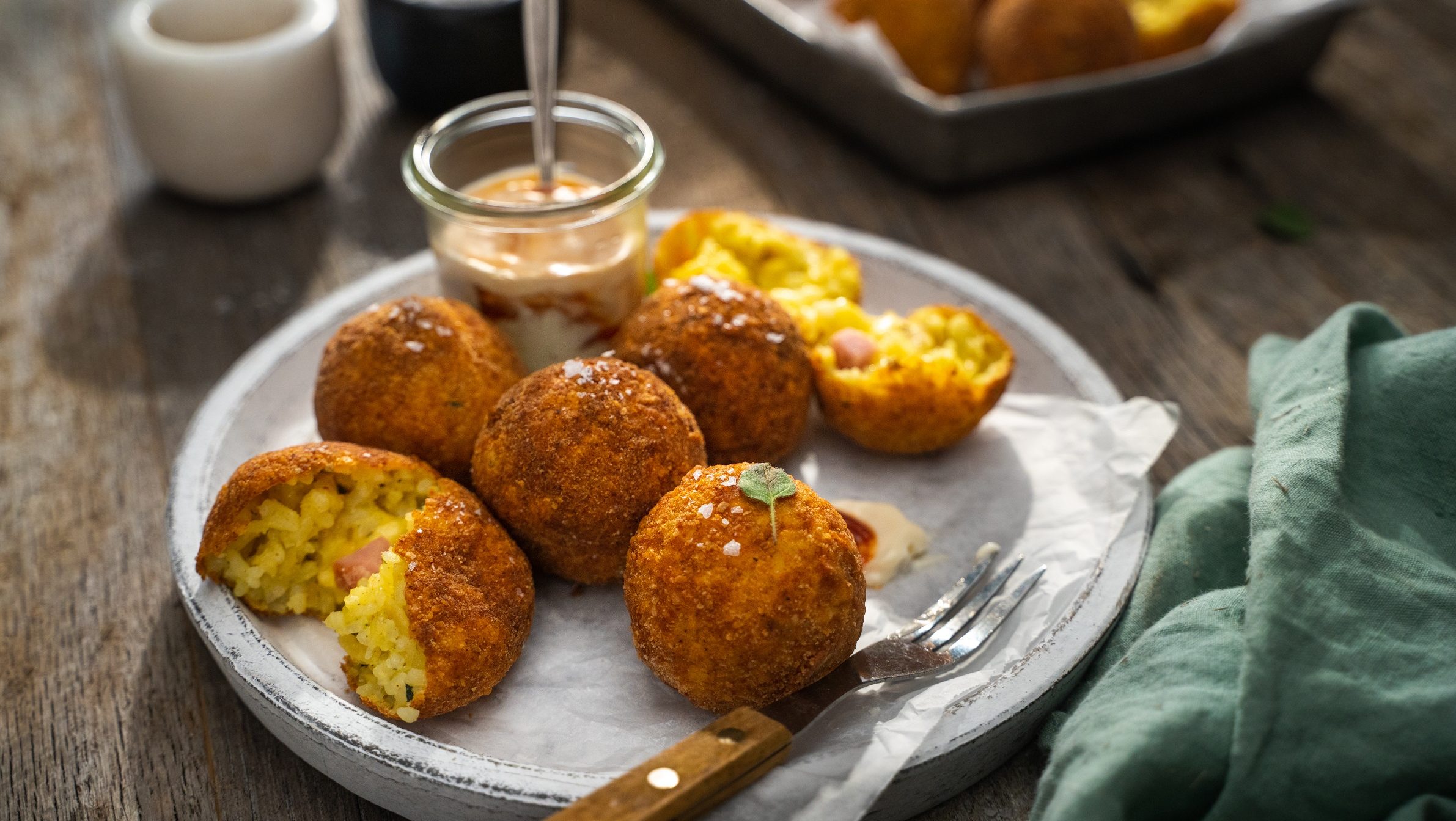 Five ham and cheese arancini balls on a white plate with a fork and a pot of sauce on a wooden table.