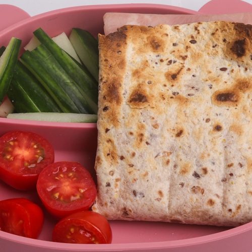 A toasted folded wrap, cucumber sticks and cherry tomatoes in a teddy bear shaped pink lunchbox.
