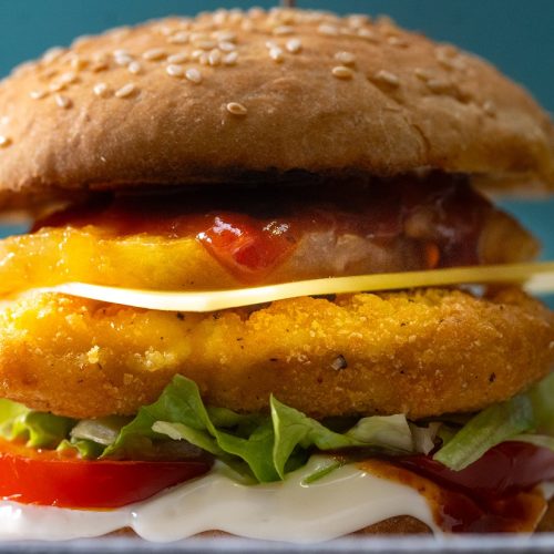 A side view of a crumbed chicken burger with tomato, lettuce, cheese, pineapple and red sauce between buns.